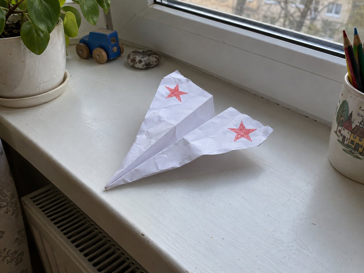 Бумажный самолётик из белого листа с острым носом и отогнутыми крыльями, на крыльях нарисованы красные звёзды